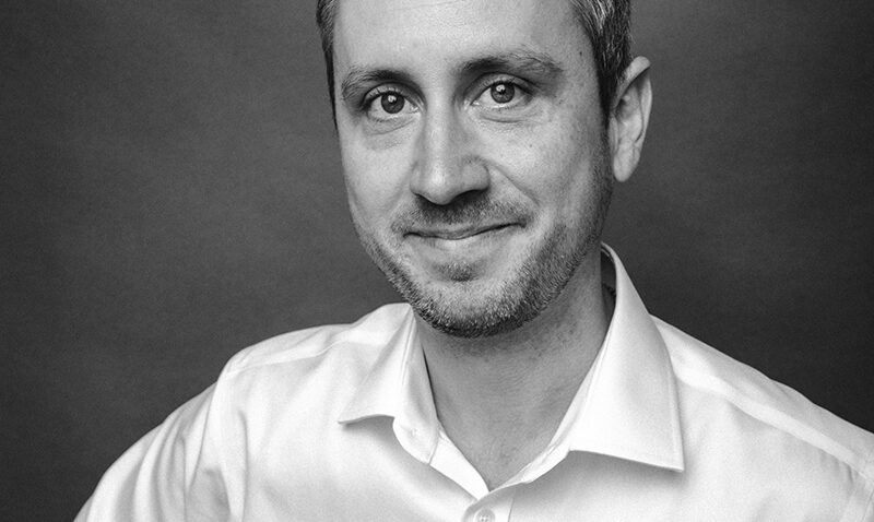 Christopher Nairne, a man with short hair and light stubble, wearing a white button-up shirt, smiles slightly whilst looking at the camera. The photo is in black and white with a plain dark background.