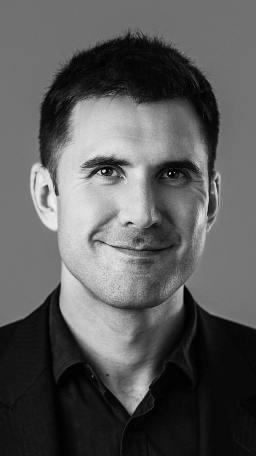 Christian Barraclough, a man with short dark hair, wearing a dark shirt and blazer, smiles softly against a plain grey background in this black-and-white portrait.