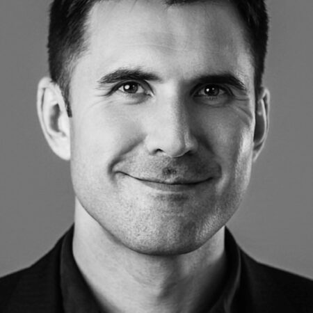 Christian Barraclough, a man with short dark hair, wearing a dark shirt and blazer, smiles softly against a plain grey background in this black-and-white portrait.