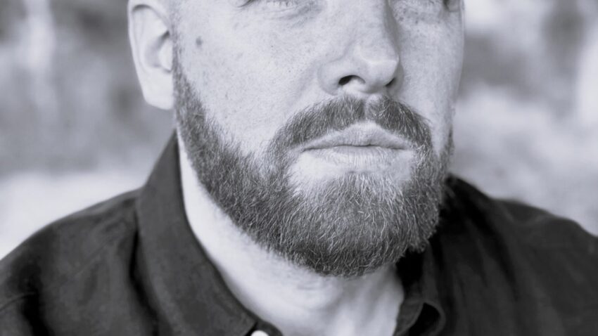 Black and white portrait of David Shipley, a bearded man with short hair, wearing a dark button-up shirt, looking directly at the camera with a serious expression. Blurred outdoor background.