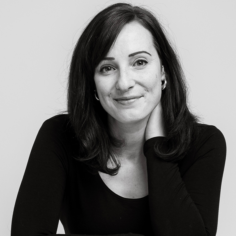 Eleanor Pennell-Briggs, with shoulder-length dark hair and a black long-sleeved top, sits with her hand resting on her neck and smiles gently at the camera in this black-and-white photo.