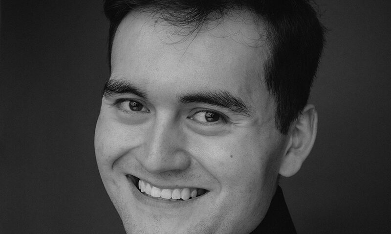 A young man with dark hair smiles warmly at the camera. Philippe Durrant is wearing a dark shirt and is posed against a plain, dark background in this black and white portrait.