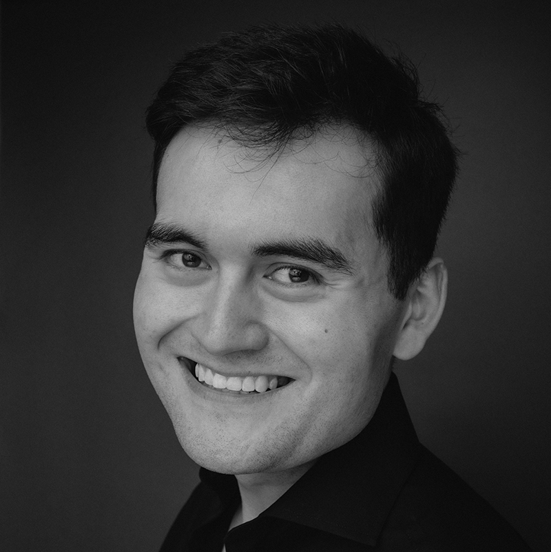 A young man with dark hair smiles warmly at the camera. Philippe Durrant is wearing a dark shirt and is posed against a plain, dark background in this black and white portrait.