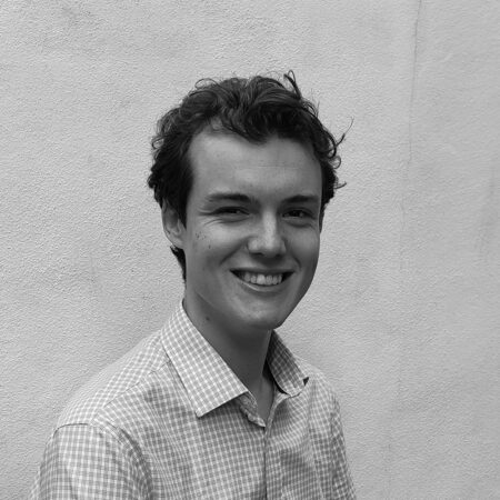 Samuel Kibble, a young person with curly hair and a chequered shirt, smiles at the camera while standing in front of a plain, textured wall. The black and white image captures his cheerful expression beautifully.