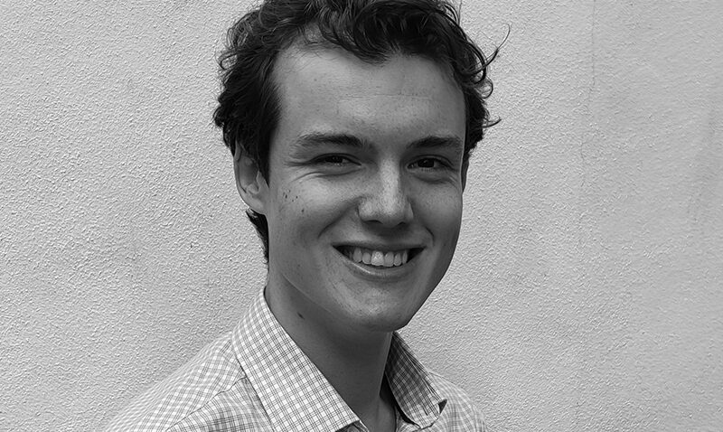 Samuel Kibble, a young person with curly hair and a chequered shirt, smiles at the camera while standing in front of a plain, textured wall. The black and white image captures his cheerful expression beautifully.