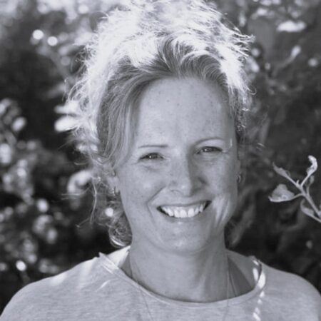 Sophia Durrant, a woman with fair hair pulled back, smiles outdoors in a light-coloured shirt. Sunlight filters through the leaves in the blurred background. The image is in black and white.