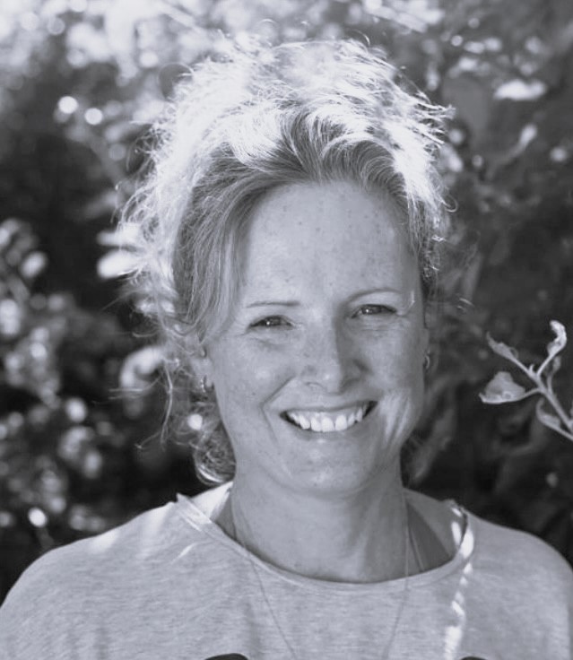 Sophia Durrant, a woman with fair hair pulled back, smiles outdoors in a light-coloured shirt. Sunlight filters through the leaves in the blurred background. The image is in black and white.