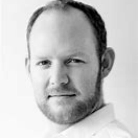 A man with a beard and short hair, wearing a collared shirt, looks directly at the camera with a neutral expression against a plain, light background. This portrait captures Dominic J. Walsh in a moment of calm composure.