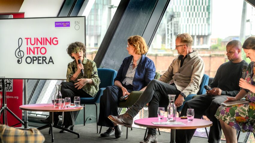 Five people sit on a stage in discussion, with one person speaking into a microphone. A large screen next to them displays the text TUNING INTO OPERA, highlighting a lively conversation about music and Opera. Glasses of water rest on small tables nearby.