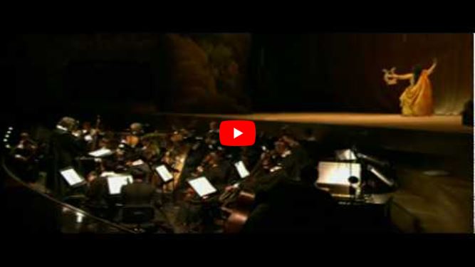 An orchestra plays in a dimly lit pit, evoking the world of Orpheus, whilst a dancer in a yellow dress performs under a spotlight on stage.