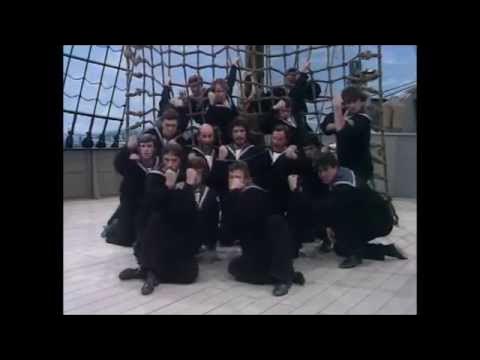 Screenshot from A British Tar from HMS Pinafore. Image of a group of men dressed as sailors pose dramatically on the deck of a ship, with some kneeling and others standing in front of a rope net on the mast—capturing the spirited charm found in Gilbert & Sullivan music.