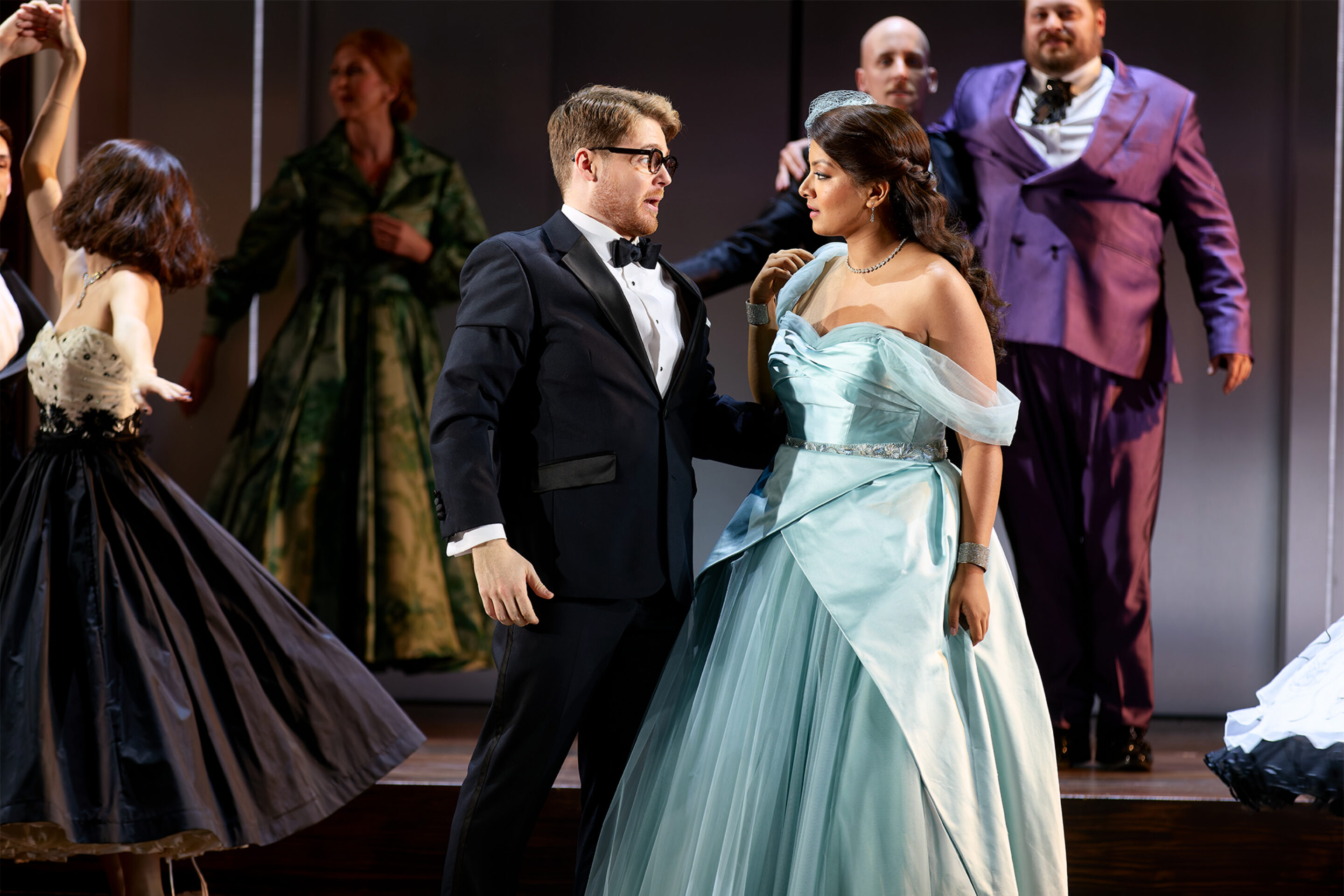 Aaron Godfrey Mayes, Deepa Johnny, ENO’s Cinderella (La Cenerentola) 2025 © Mark Douet A man in a black dinner jacket and a woman in a light blue ball gown stand together on stage, surrounded by other people in formal dress, suggesting a dramatic or theatrical performance.