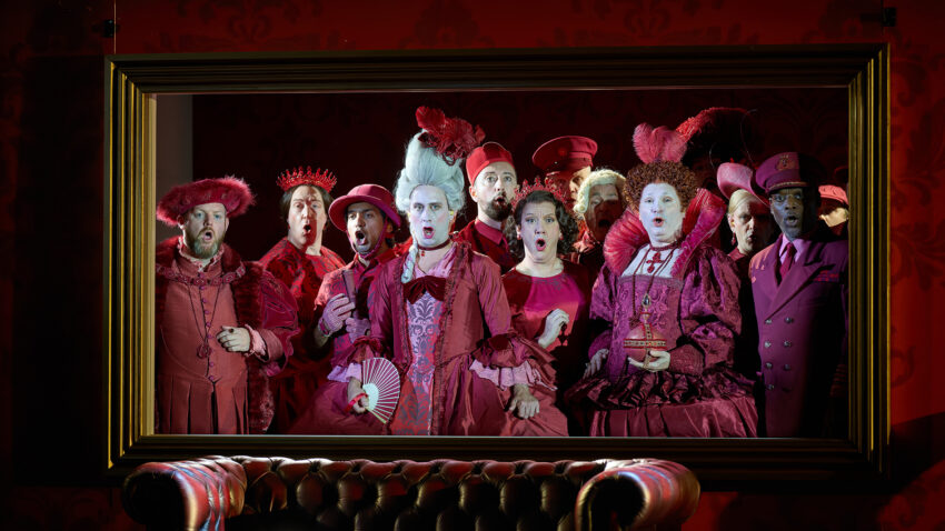 Chorus of ENO, ENO’s Cinderella (La Cenerentola) 2025 © Mark Douet A group of people in elaborate, old-fashioned costumes and white face make-up, all dressed in various shades of red, stand closely together in a gold-framed setting, looking ahead with shocked expressions.
