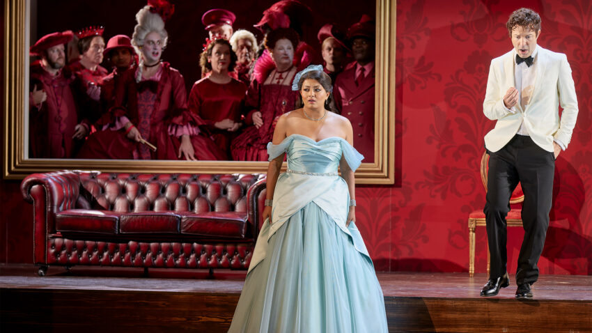 Deepa Johnny, Charles Rice, Chorus of ENO, ENO’s Cinderella (La Cenerentola) 2025 © Mark Douet A woman in a blue dress stands in front of a red settee, while a man in a white jacket sings beside her. Behind them, a large framed picture shows a group of people in elaborate red costumes and wigs.