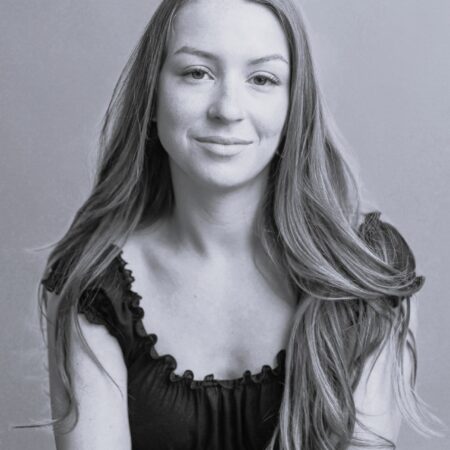 Natasha Oldbury, a young woman with long, wavy hair, sits against a plain background, looking at the camera with a gentle smile. She wears a short-sleeved, frilled black top. The photo is in black and white.