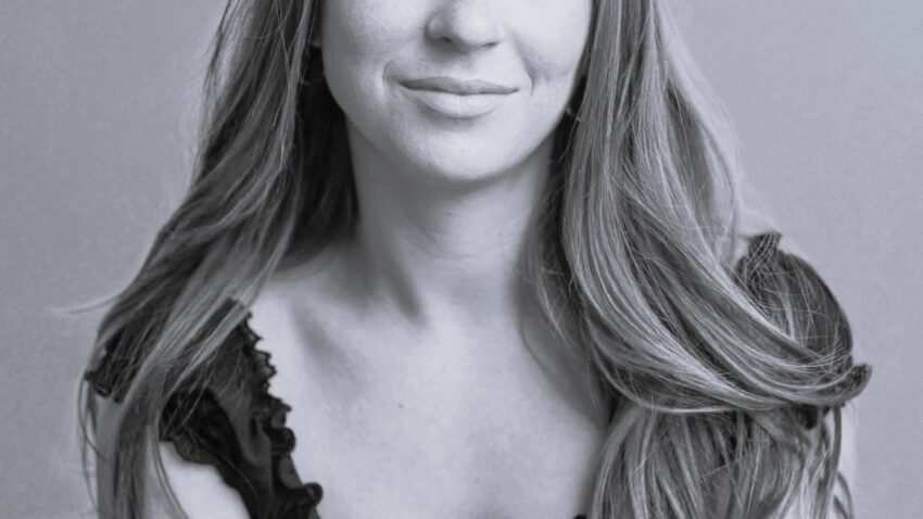 Natasha Oldbury, a young woman with long, wavy hair, sits against a plain background, looking at the camera with a gentle smile. She wears a short-sleeved, frilled black top. The photo is in black and white.