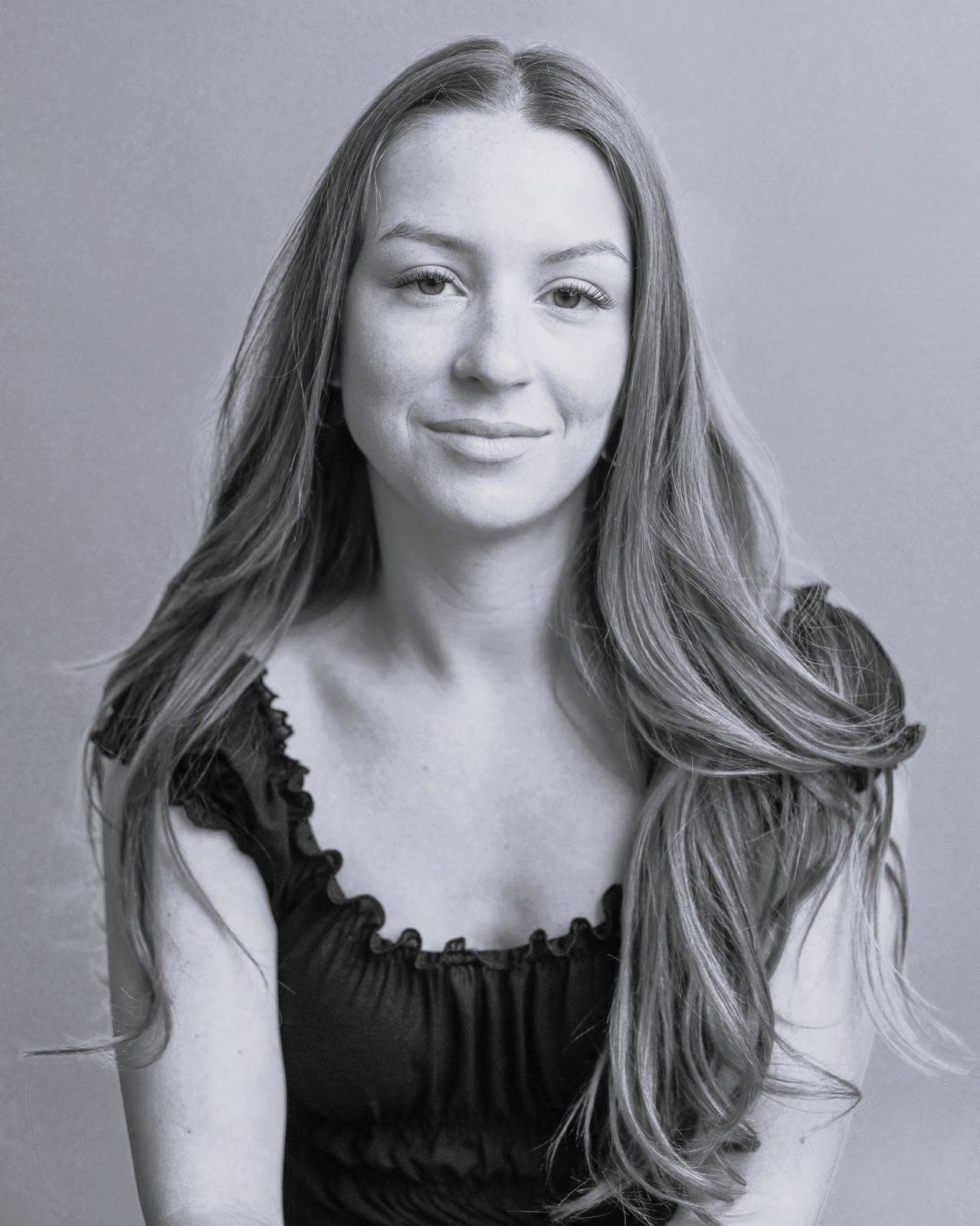 Natasha Oldbury, a young woman with long, wavy hair, sits against a plain background, looking at the camera with a gentle smile. She wears a short-sleeved, frilled black top. The photo is in black and white.