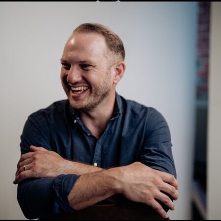 A man with short hair and a beard, wearing a dark blue button-up shirt, smiles and laughs with his arms crossed, sitting against a plain, light-coloured background.