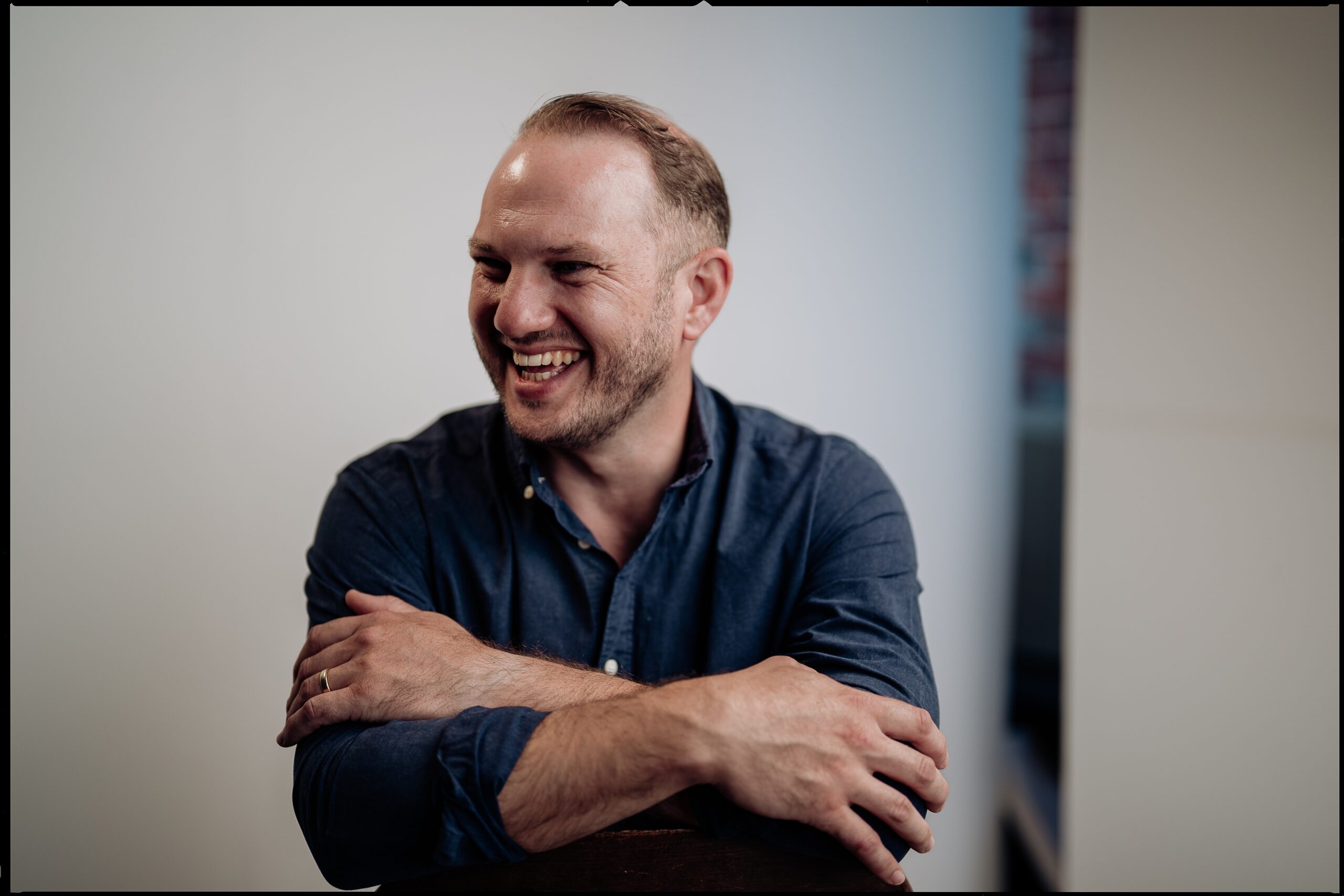 A man with short hair and a beard, wearing a dark blue button-up shirt, smiles and laughs with his arms crossed, sitting against a plain, light-coloured background.
