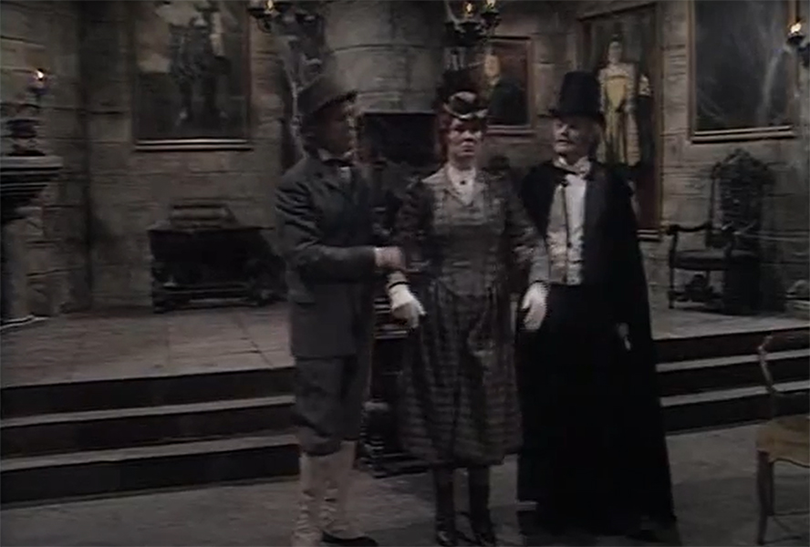 Three people in Victorian-era costumes stand in a dimly lit, old-fashioned room with stone walls, paintings, and a suit of armour—reminiscent of a scene from one of Gilbert & Sullivan's best songs.