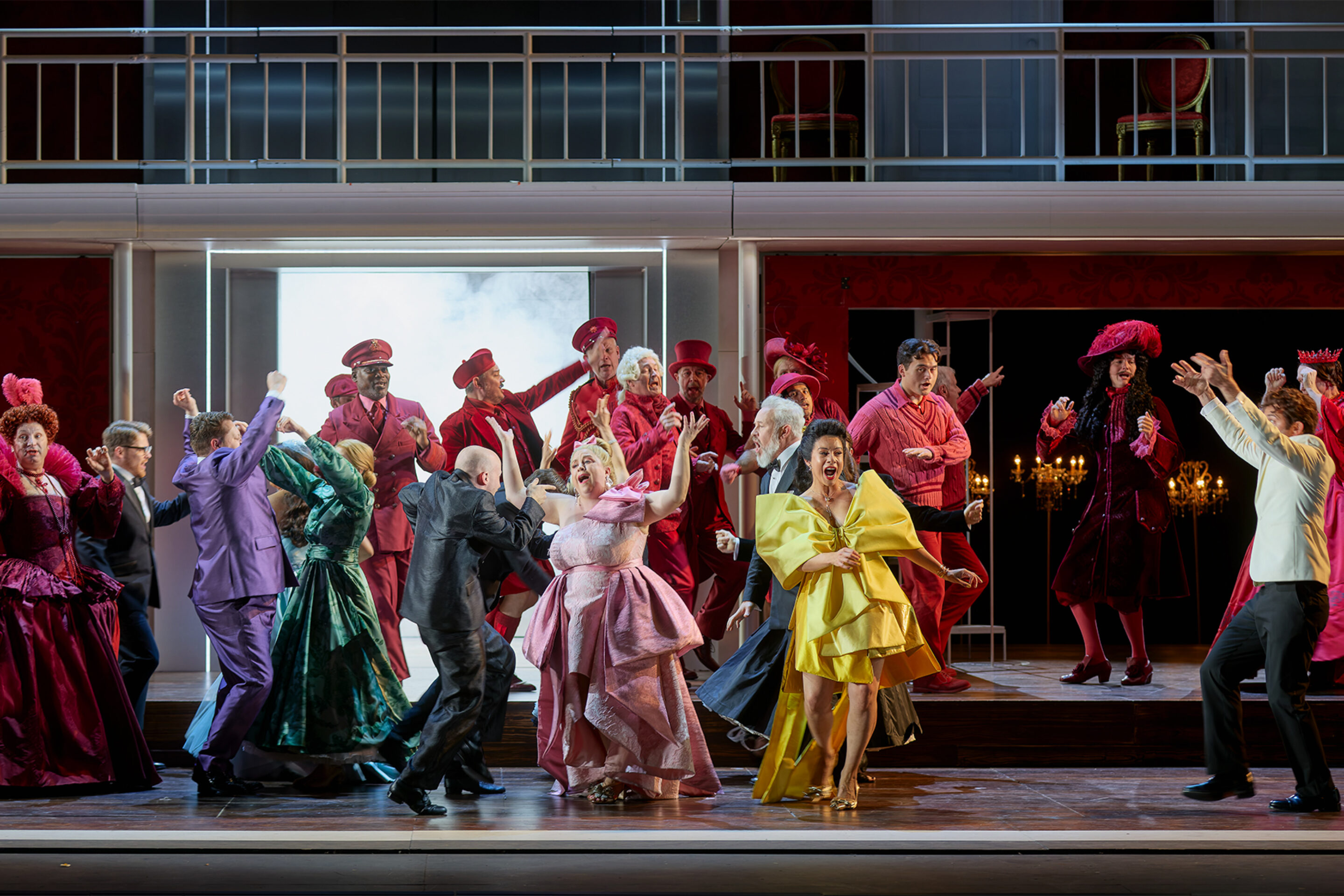 The Cast of ENO’s Cinderella (La Cenerentola) 2025 © Mark Douet A lively stage scene with actors dressed in colourful costumes—mostly reds, pinks, and yellows—dancing and gesturing energetically, set against an elegant, multi-level backdrop with chandeliers and dramatic lighting.
