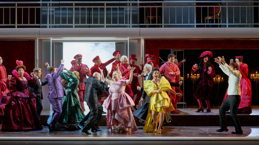 The Cast of ENO’s Cinderella (La Cenerentola) 2025 © Mark Douet A lively stage scene with actors dressed in colourful costumes—mostly reds, pinks, and yellows—dancing and gesturing energetically, set against an elegant, multi-level backdrop with chandeliers and dramatic lighting.