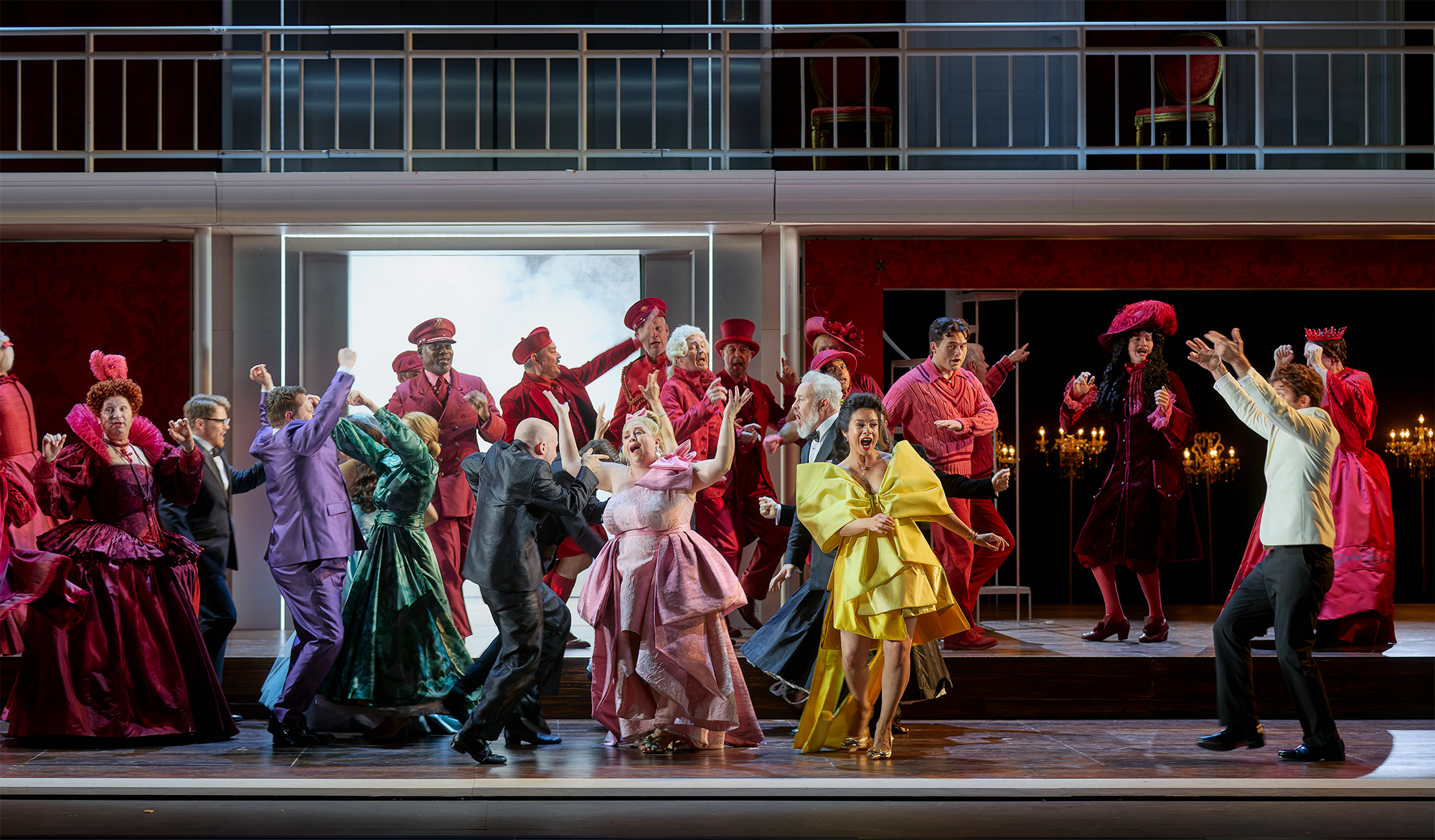 The Cast of ENO’s Cinderella (La Cenerentola) 2025 © Mark Douet A lively stage scene with actors dressed in colourful costumes—mostly reds, pinks, and yellows—dancing and gesturing energetically, set against an elegant, multi-level backdrop with chandeliers and dramatic lighting.