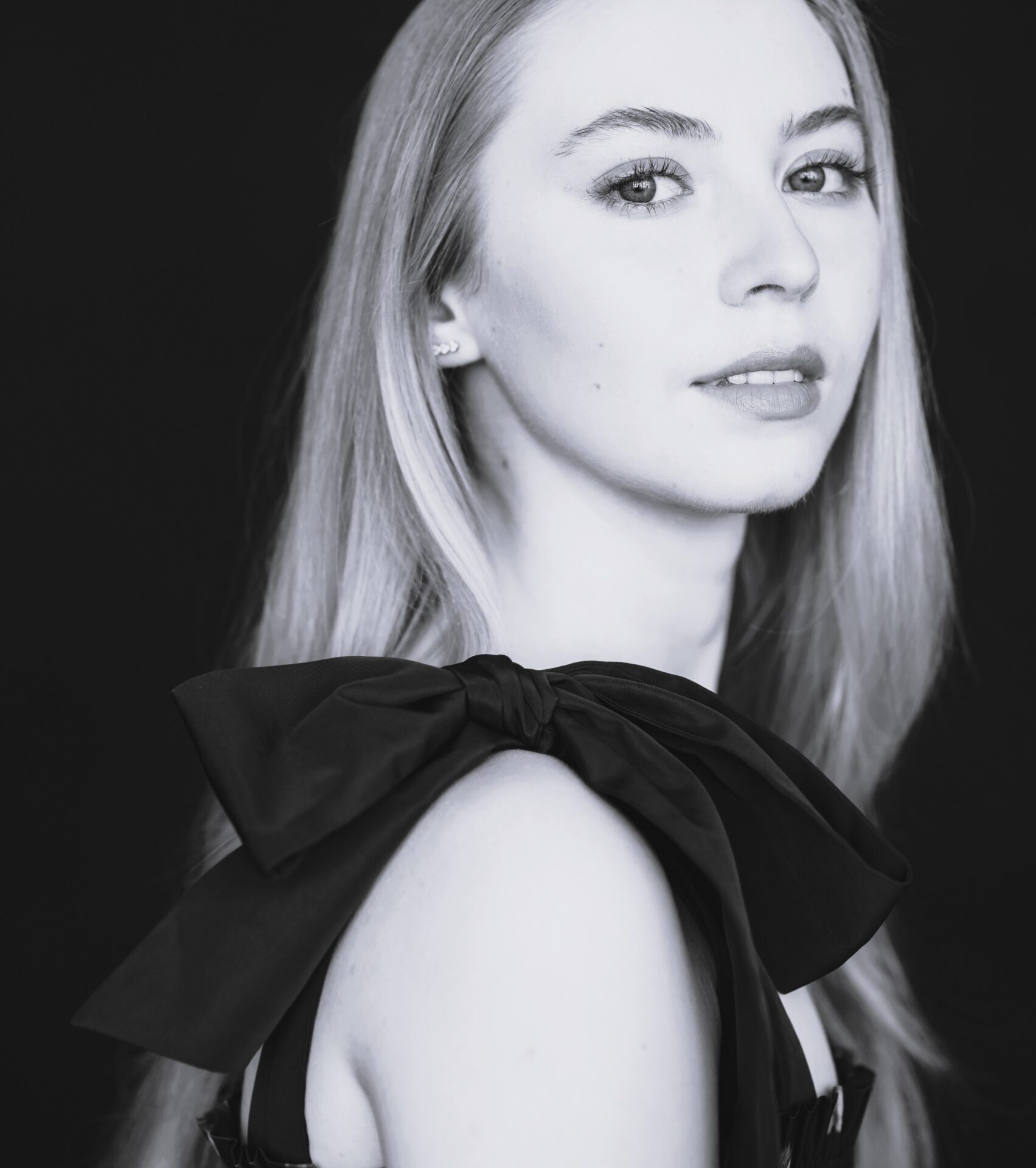 Black and white portrait of Abigail Sinclair, a young woman with long, straight hair, wearing a sleeveless dress with a large bow on the shoulder, looking slightly over her shoulder at the camera against a dark background.