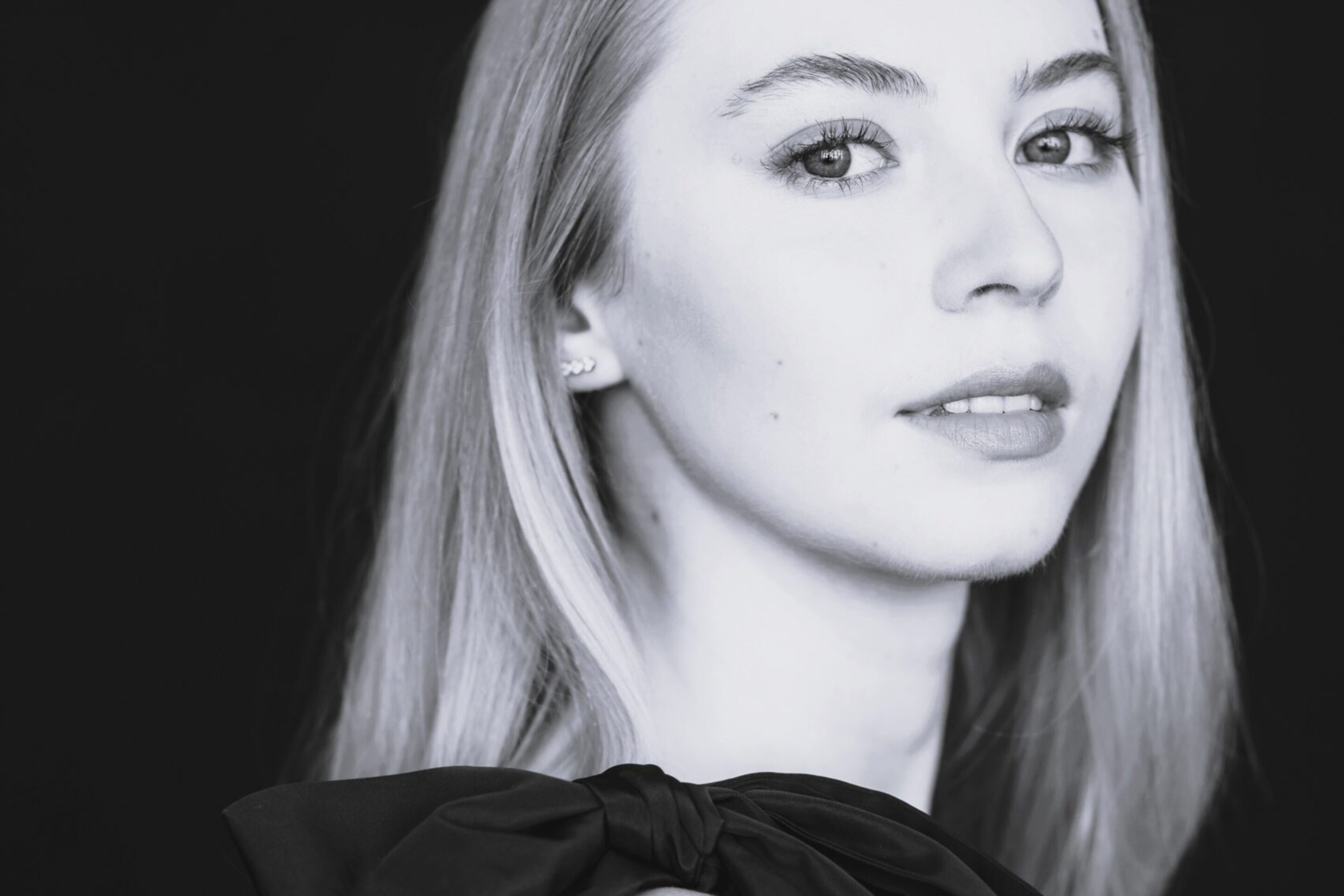 Black and white portrait of Abigail Sinclair, a young woman with long, straight hair, wearing a sleeveless dress with a large bow on the shoulder, looking slightly over her shoulder at the camera against a dark background.