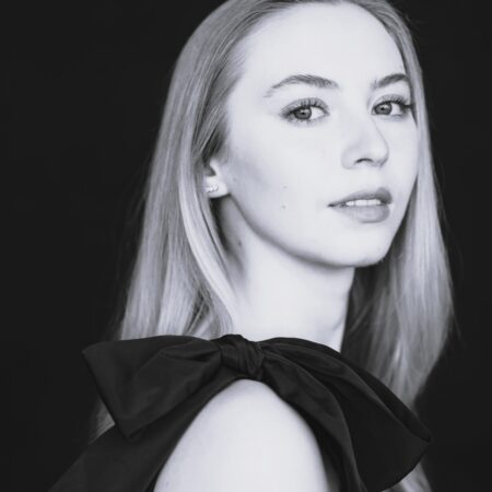 Black and white portrait of Abigail Sinclair, a young woman with long, straight hair, wearing a sleeveless dress with a large bow on the shoulder, looking slightly over her shoulder at the camera against a dark background.