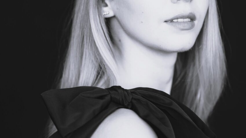 Black and white portrait of Abigail Sinclair, a young woman with long, straight hair, wearing a sleeveless dress with a large bow on the shoulder, looking slightly over her shoulder at the camera against a dark background.