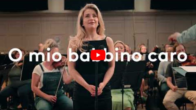 A blonde woman in a black dress stands at a microphone before an orchestra, performing one of opera’s iconic roles. Large white text across the image reads O mio babbino caro. A conductor’s hand is visible on the right.