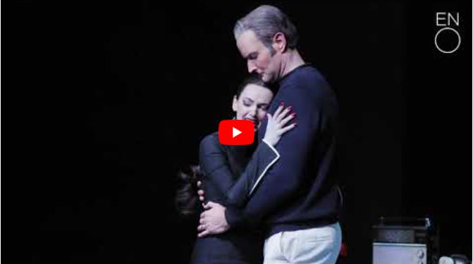 A woman in black embraces a tall man in dark clothing onstage, both with serious expressions, evoking the timeless story of Orpheus.
