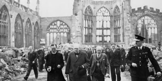 A group of men, including officials and clergy, walk through the ruins of a bombed-out cathedral.