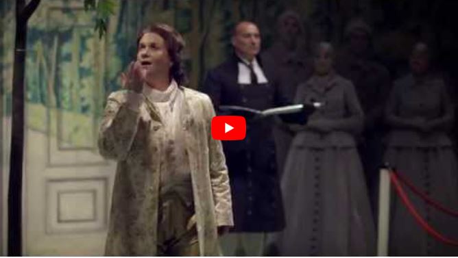 A man in a light-coloured period costume stands in the foreground, gesturing while singing.