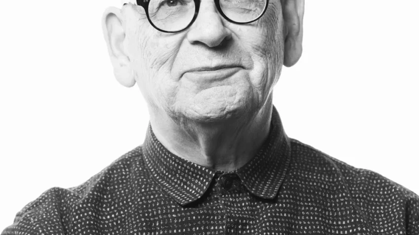 Black and white portrait of Anthony McDonald, an older man with glasses, a bald head, and a slight smile. He is wearing a patterned button-up shirt and facing the camera against a plain, light background.