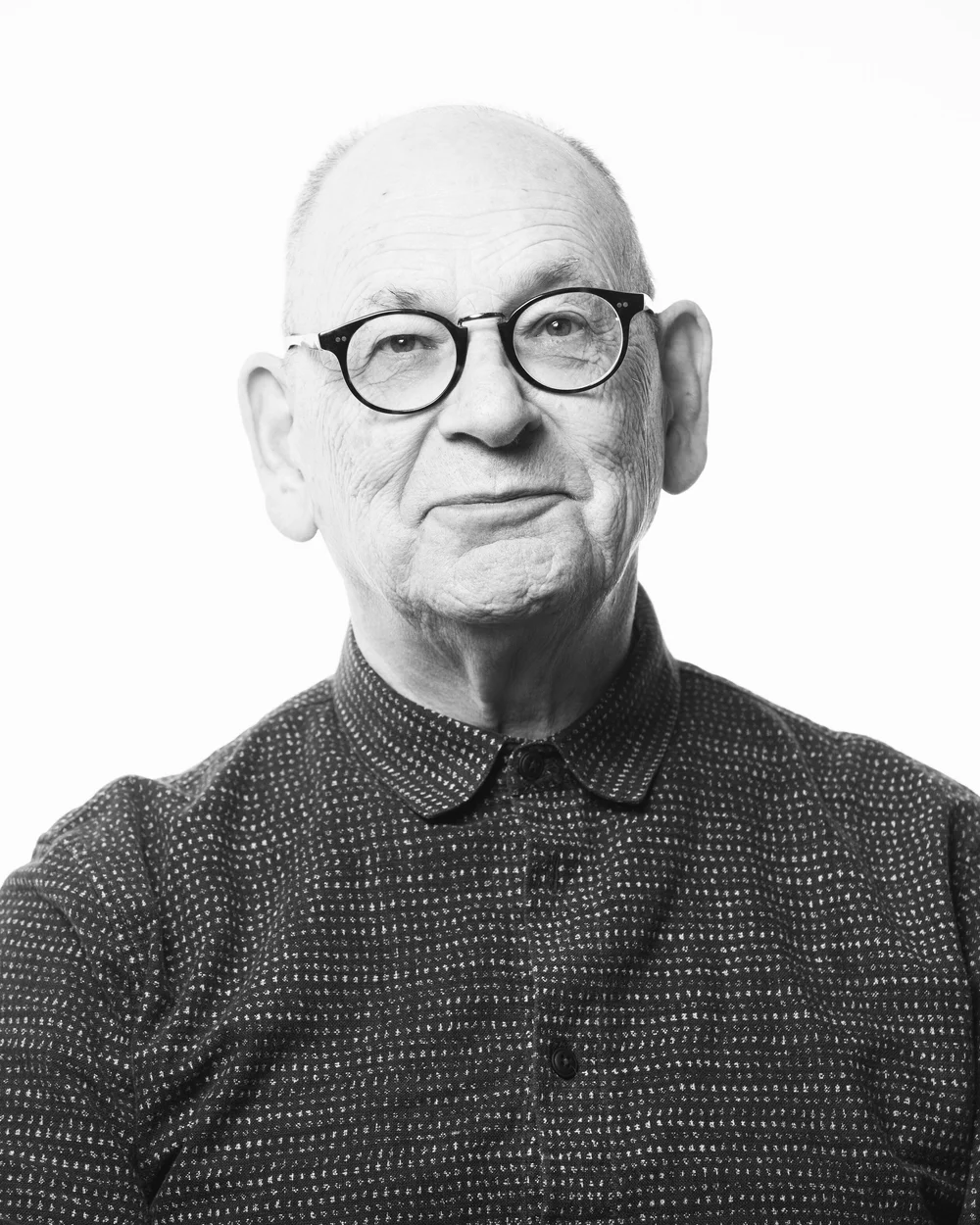 Black and white portrait of Anthony McDonald, an older man with glasses, a bald head, and a slight smile. He is wearing a patterned button-up shirt and facing the camera against a plain, light background.