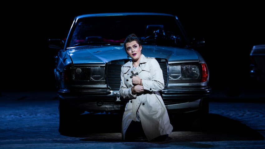Ava Dodd, ENO’s Carmen 2025 © Ellie Kurttz A woman in a light trench coat kneels on the ground in front of an old car at night, clutching her coat and looking upwards with a worried expression. Dramatic stage lighting highlights her and the vehicle.