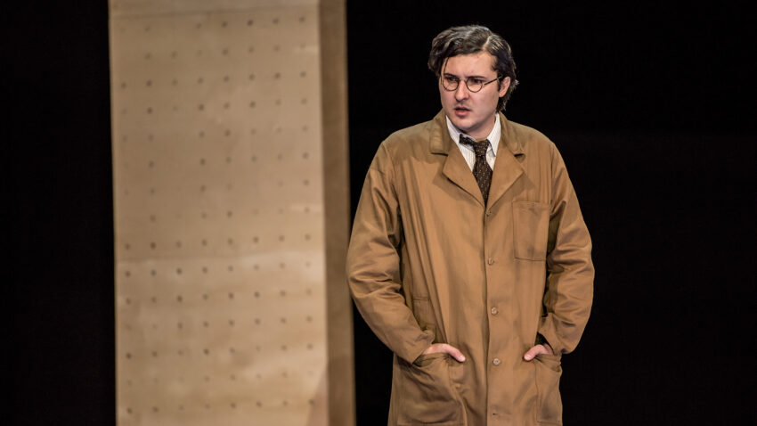 A person wearing glasses, a brown coat, and a tie stands with hands in pockets in front of a light wooden wall with holes, appearing to be on a stage set.