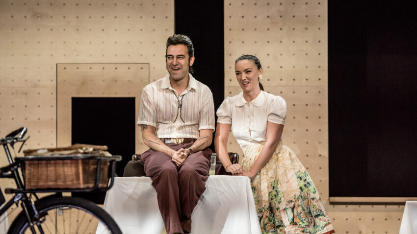 A man and a woman in vintage-style clothing sit side by side on a stage, smiling. The set features a bicycle with a basket in the foreground and simple, light-coloured walls with black rectangular shapes.