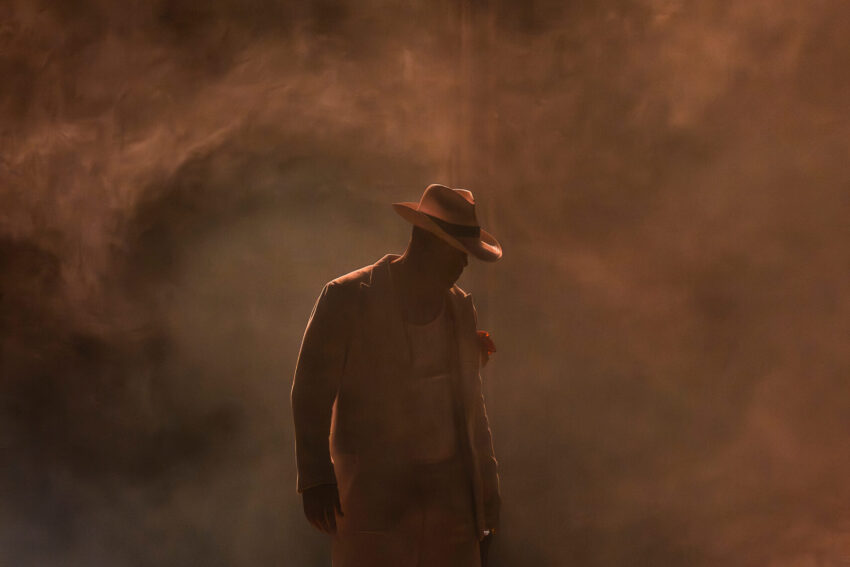Dean Street, ENO’s Carmen 2025 © Ellie Kurttz A person wearing a suit and hat stands onstage under dramatic lighting, surrounded by swirling smoke or fog, creating a mysterious and atmospheric scene.
