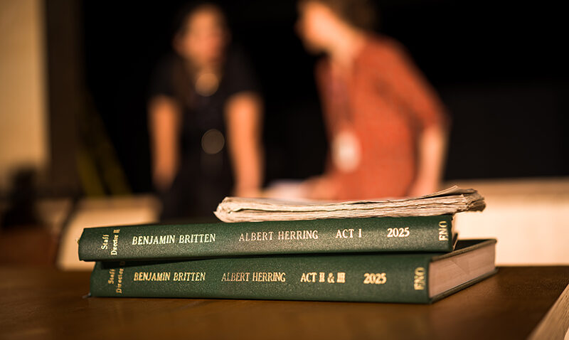 Two green bound scripts labelled Benjamin Britten: Albert Herring, Act I & II sit on a table with papers on top; in the blurred background at the Lowry, Greater Manchester, two people sit and talk.