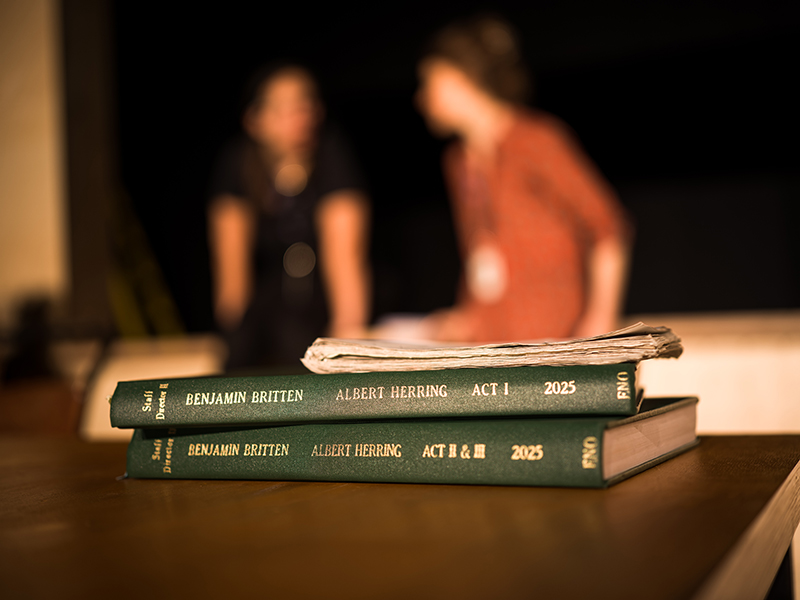 Two green bound scripts labelled Benjamin Britten: Albert Herring, Act I & II sit on a table with papers on top; in the blurred background at the Lowry, Greater Manchester, two people sit and talk.