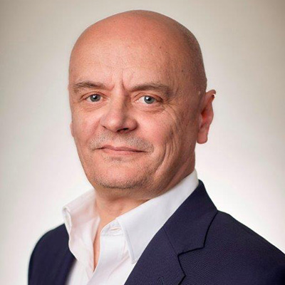 ENO & LCL Board Member Simon Bailey wearing a dark suit jacket and a white dress shirt, smiles slightly whilst posing against a plain, light-coloured background, reflecting his professional role with ENO and board appointments at the London Coliseum.