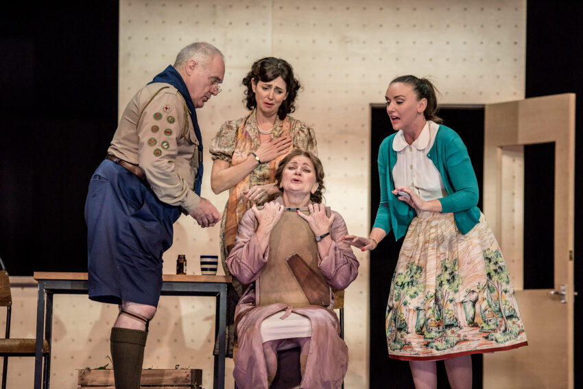 Four adults in vintage clothing perform a dramatic scene on stage. One man in a scout uniform and three women in 1950s-style outfits are gathered around a seated woman who appears distressed or surprised.