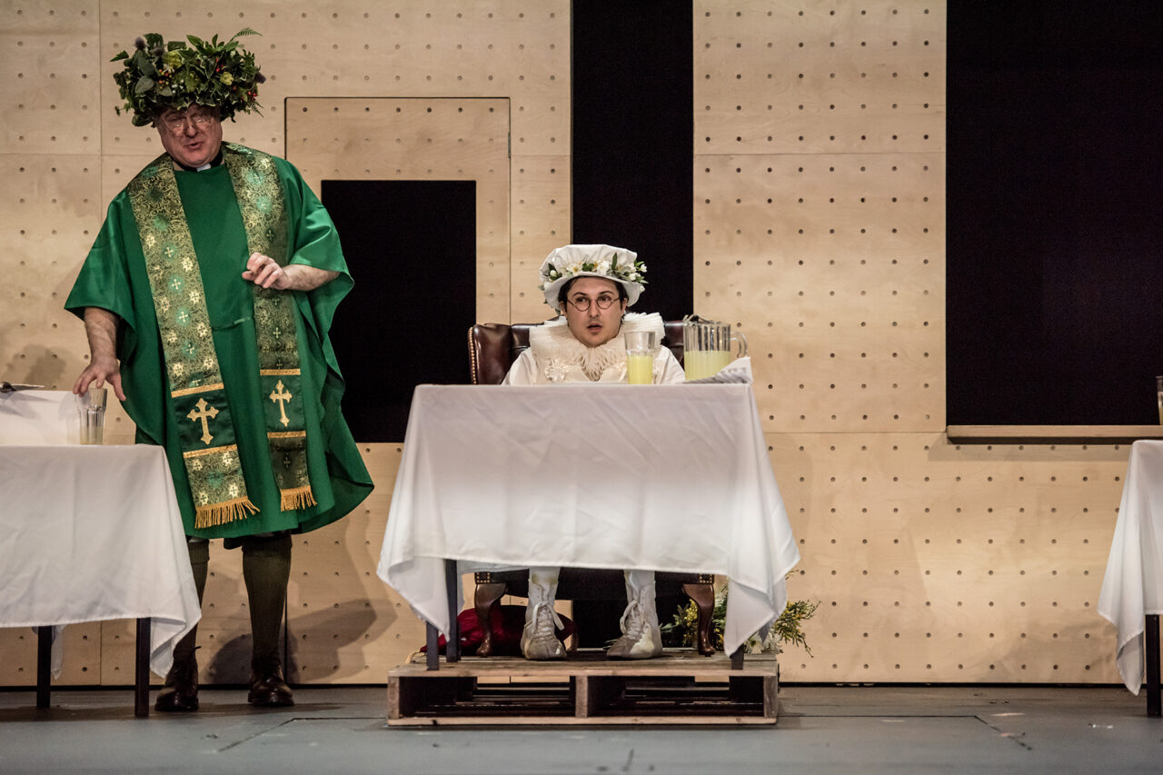 Two people in costume on stage: one standing in a green robe and leafy crown, the other sitting at a table in white attire and a hat, looking surprised. The scene appears to be part of a theatrical performance.