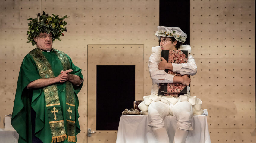 Two people in theatrical costumes perform on stage. The person on the left wears a green robe and a leafy crown, while the person on the right, dressed in white with a chef’s hat, sits clutching a large book and looks at the other.