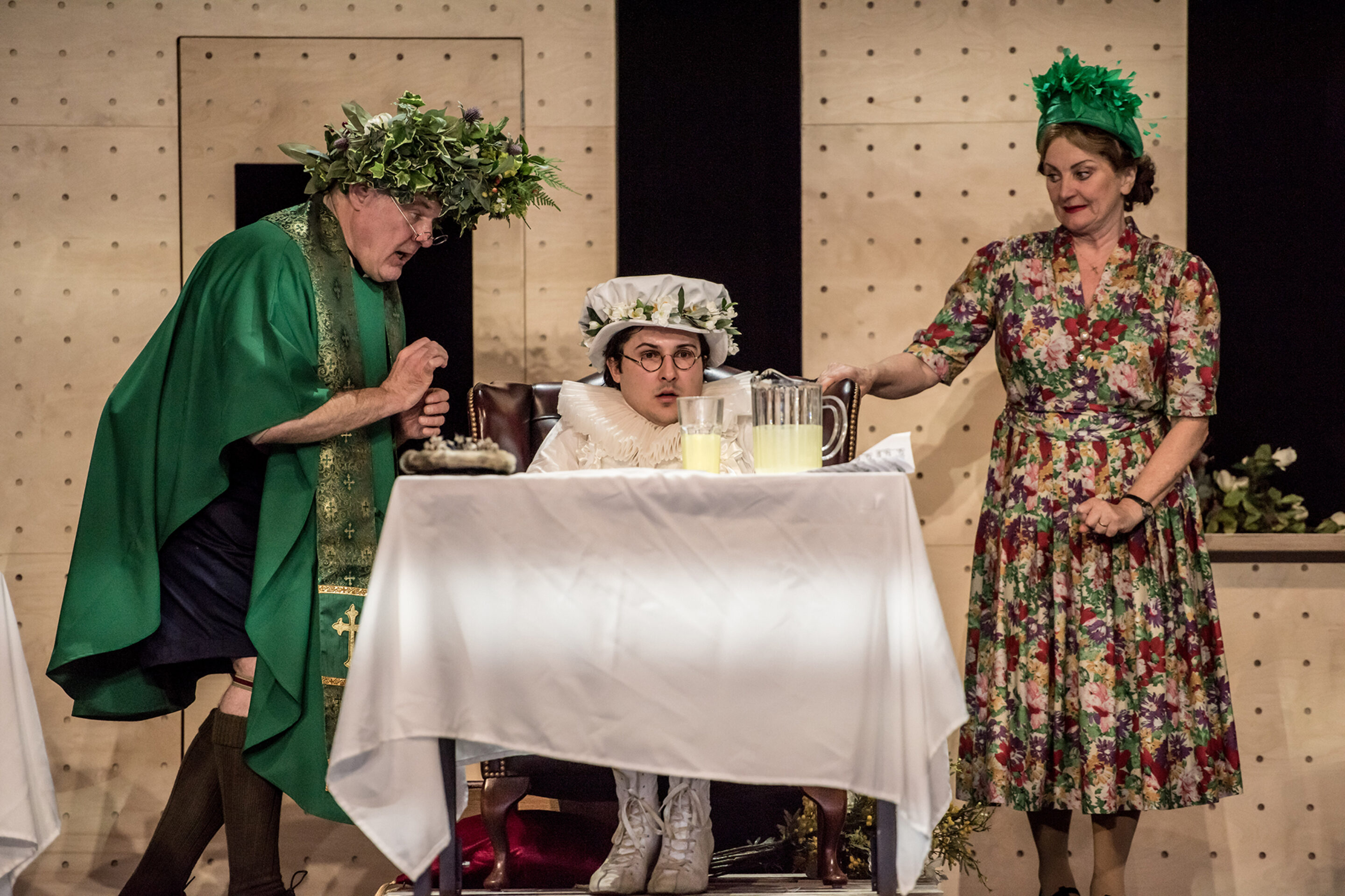 Three people in theatrical costumes are on stage. One, in green robe and leaf crown, stands beside another seated in white clothes and hat. A woman in a flowered dress and green headpiece pours lemonade at a table.