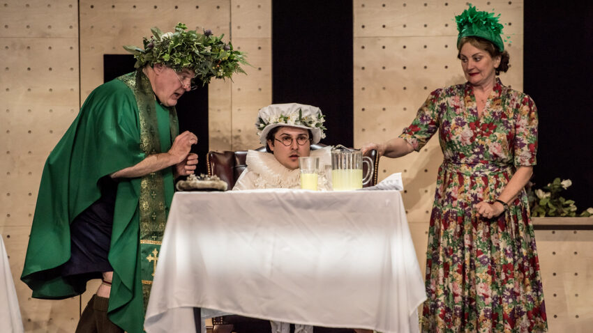 Three people in theatrical costumes are on stage. One, in green robe and leaf crown, stands beside another seated in white clothes and hat. A woman in a flowered dress and green headpiece pours lemonade at a table.