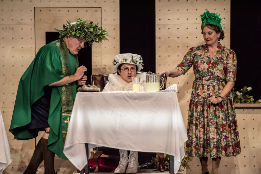 Three people in theatrical costumes are on stage. One, in green robe and leaf crown, stands beside another seated in white clothes and hat. A woman in a flowered dress and green headpiece pours lemonade at a table.